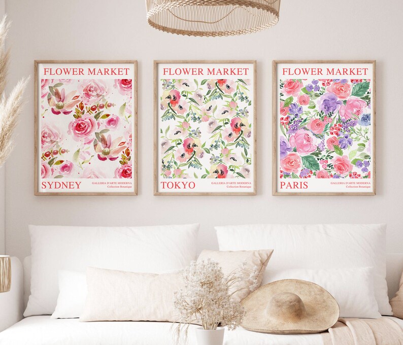 May include: Three framed floral art prints with the text "Flower Market" above the city names Sydney, Tokyo, and Paris. The prints feature watercolor floral patterns in pink, purple, and green hues. The prints are displayed above a white sofa.