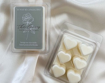 May include: Six heart-shaped wax melts in a clear plastic container. The wax melts are white and have a floral scent called "The Lovers" with oud, patchouli, and resin notes. The container is labeled "Luna Fae Collective" and "Hemp Soy Wax Melts, Hand Poured in Florida."