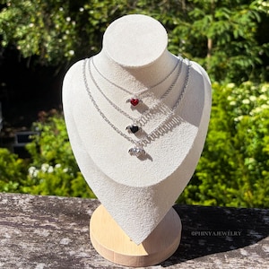 Könnte beinhalten: Drei mehrreihige Halsketten mit Herzanhängern auf einer weißen Schmuckausstellung. Jede Halskette hat ein andersfarbiges Herz: rot, schwarz und silber. Die Herzen haben kleine Flügeldetails. Der Hintergrund ist ein verschwommenes Grün.