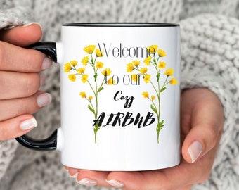 May include: White ceramic mug with a black handle and rim, featuring the text "Welcome to our Cozy AIRBNB" with yellow floral accents. The mug is held by a person wearing a gray cable-knit sweater.