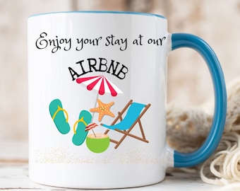 May include: White ceramic mug with a blue rim and handle. The mug has a graphic of a beach scene with an umbrella, flip flops, a lounge chair, a coconut drink, and a starfish. The text "Enjoy your stay at our AIRBNB" is printed on the mug.