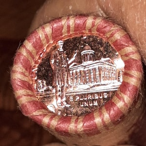 May include: A roll of U.S. coins, with a visible coin featuring a man and a building. The roll is wrapped in red and tan striped paper. The coin has the inscription "E PLURIBUS UNUM".