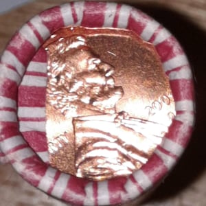 May include: A roll of pennies, wrapped in a red and white striped paper. The copper-colored coins are visible at the end of the roll, with the profile of Abraham Lincoln facing left. The year 2000 is visible on the coin.