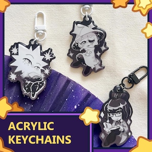 May include: Three acrylic keychains featuring black and white anime-style character illustrations. One keychain shows a spiky-haired figure, another a character in a wide-brimmed hat, and the third a figure with glasses reading a book. Each has a clasp.