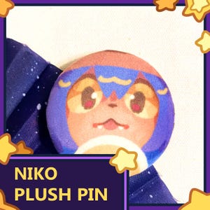 May include: A round plush pin featuring a cartoon character with orange, brown, and blue colors. The pin has a smiling face with large eyes. The text "NIKO PLUSH PIN" is displayed on a purple background below the pin. The background is white and blue.