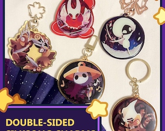 May include: A collection of double-sided Silksong charms featuring various character designs. The charms are round and have gold-colored keychains. The designs include characters in red, white, purple, and brown colors. The text "DOUBLE-SIDED SILKSONG CHARMS" is visible.