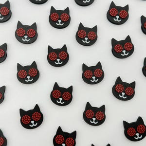 May include: A collection of black cat-shaped buttons with red rhinestone eyes. The buttons are arranged in a staggered pattern on a white background.