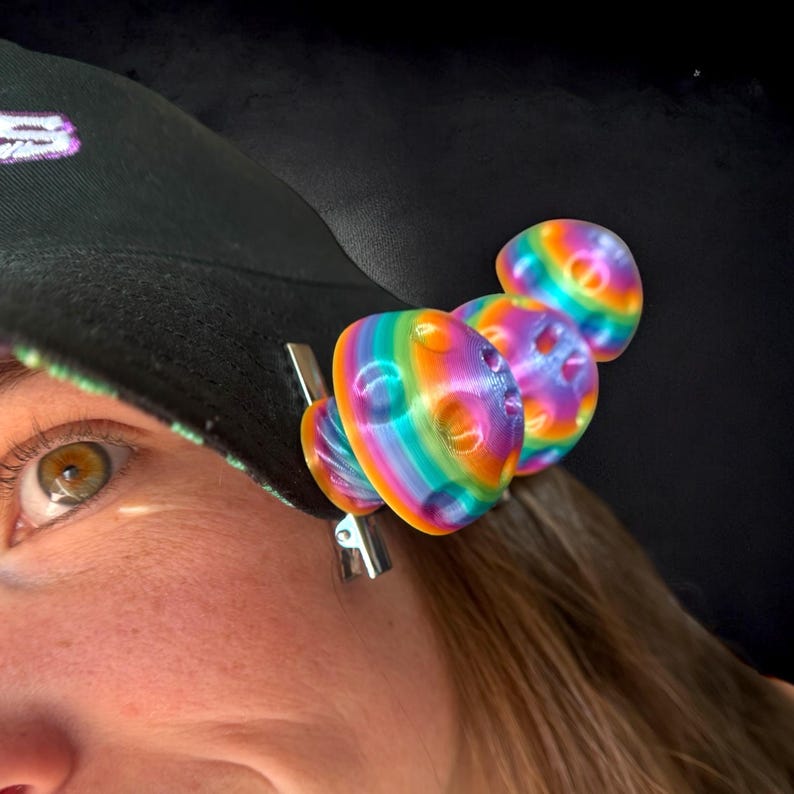 May include: A black baseball cap with a rainbow-colored clip-on accessory. The accessory has a series of three rounded shapes with a striped pattern of multiple colors. The clip is silver.
