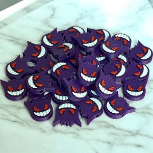 May include: A pile of purple and white plastic charms shaped like the Pokemon character, Gengar. The charms have red eyes and a white smile.