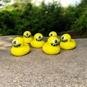 May include: A group of six yellow rubber duck toys with a single large eye and a smiling mouth. The ducks are arranged on a concrete surface with a blurred green background of foliage.