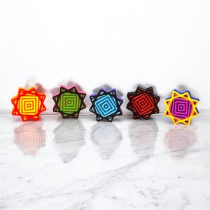 May include: Five colorful star-shaped ornaments with geometric designs. The ornaments are red, green, blue, black, and pink.