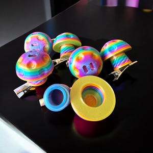 May include: A collection of colorful, rainbow-striped mushroom-shaped hair clips and accessories. The clips have a vibrant, multi-colored design with a metallic silver clip. Other accessories include a blue and yellow ring-shaped object.