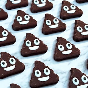 May include: Rows of chocolate candies shaped like smiling poop emojis. Each dark brown candy has white eyes and a white smiling mouth. The candies are arranged on a white and gray marble surface.