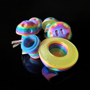 May include: A collection of colorful, rainbow-striped 3D-printed objects. The objects include mushroom-shaped items, a ring, and a spiral-shaped piece. The items are set against a black background, with reflections visible.