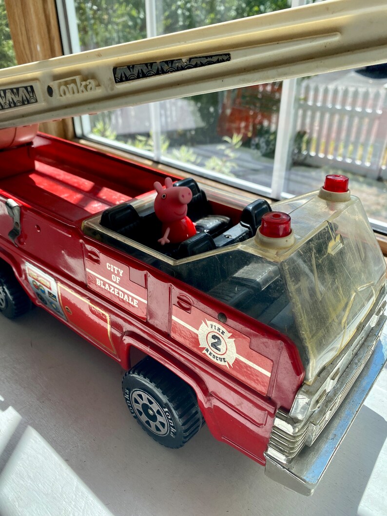 May include: A red toy fire truck with the text "City of Blazedale" and "Fire 2 Rescue". A pink pig figurine is in the driver's seat. The truck has a white ladder and black tires.