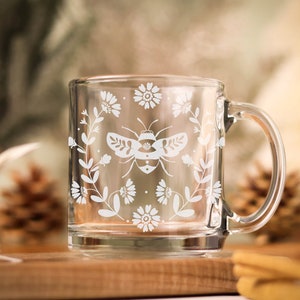 May include: A clear glass mug with a white floral and bee design. The mug has a handle and is sitting on a wooden surface.