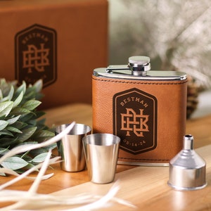 May include: A brown leather-wrapped flask with a silver top and a silver funnel. The flask is engraved with the text "Best Man" and a monogram. Two silver shot glasses are also included.