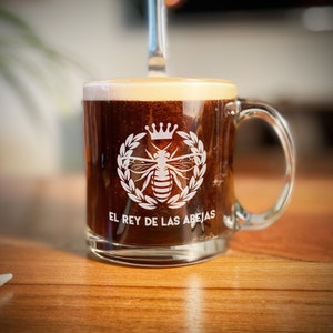 May include: A clear glass coffee mug with a white bee and crown design. The mug reads "El Rey de las Abejas".