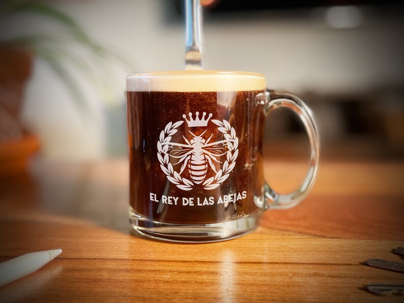 May include: A clear glass coffee mug with a white bee and crown design. The mug reads "El Rey de las Abejas".