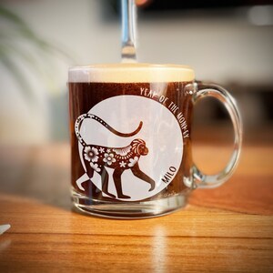 May include: A clear glass mug with a white design of a monkey with floral patterns and the text "Year of the Monkey" and "Milo". The mug is filled with coffee.
