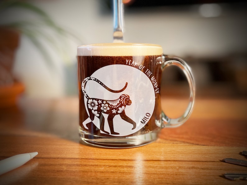 May include: A clear glass mug with a white design of a monkey with floral patterns and the text "Year of the Monkey" and "Milo". The mug is filled with coffee.
