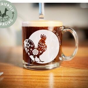 May include: A clear glass mug with a brown liquid inside. The mug has a white rooster design with the text "Year of the Rooster" on it.