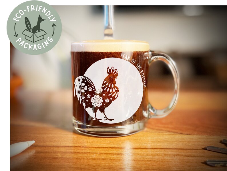 May include: A clear glass mug with a brown liquid inside. The mug has a white rooster design with the text "Year of the Rooster" on it.