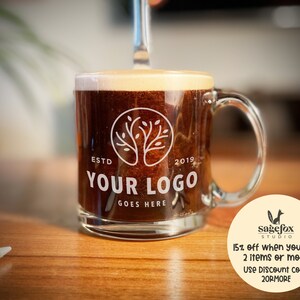 May include: A clear glass coffee mug with a brown liquid inside. The mug has a white logo with a tree design and the text "ESTD 2019 YOUR LOGO GOES HERE".