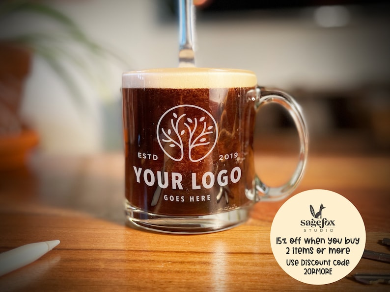 May include: A clear glass coffee mug with a brown liquid inside. The mug has a white logo with a tree design and the text "ESTD 2019 YOUR LOGO GOES HERE".