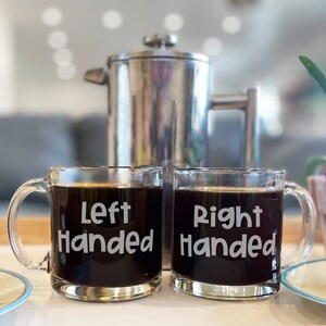 May include: Two glass coffee mugs with the words "Left Handed" and "Right Handed" printed on them. The mugs are filled with coffee and are sitting on a white surface.