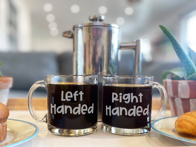 May include: Two glass coffee mugs with the words "Left Handed" and "Right Handed" printed on them. The mugs are filled with coffee and are sitting on a white surface.