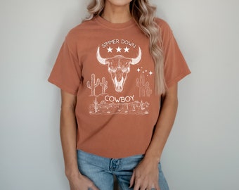 May include: A rust-colored t-shirt with a white graphic design of a bull skull, cacti, and the text "Simmer Down Cowboy".