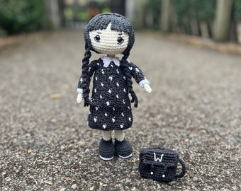 May include: A black and white crocheted doll with black hair and a white polka dot dress. The doll is holding a black crocheted bag with a white "W" on it.