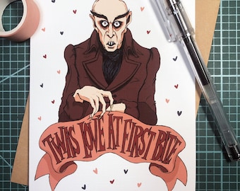 May include: A greeting card with a cartoon illustration of a vampire with a red, brown, and black outfit. The vampire is holding a banner that says "Twas love at first bite". The background is white with scattered red, pink, and gray hearts.