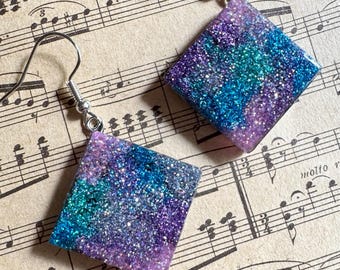 May include: A pair of square dangle earrings with a galaxy-inspired design. The earrings feature a mix of purple, teal, and blue glitter suspended in clear resin, with silver-colored fish hook ear wires. The earrings are set against a backdrop of vintage sheet music.