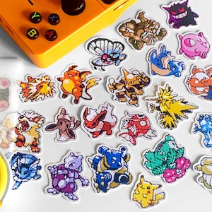 Pu&ograve; includere: Una collezione di adesivi colorati di personaggi Pok&eacute;mon pixelati, tra cui Pikachu, Charizard e Mewtwo, sono sparsi su una superficie bianca. Una console di gioco portatile gialla &egrave; sullo sfondo.