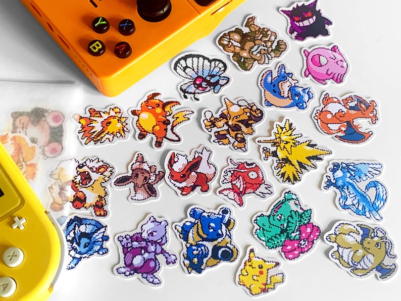 Pu&ograve; includere: Una collezione di adesivi colorati di personaggi Pok&eacute;mon pixelati, tra cui Pikachu, Charizard e Mewtwo, sono sparsi su una superficie bianca. Una console di gioco portatile gialla &egrave; sullo sfondo.