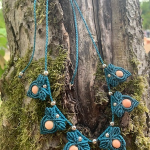 May include: Teal macrame necklace featuring a series of leaf-shaped designs, each with a peach-colored bead center. The necklace is strung with teal cord and accented with silver-colored beads. The necklace is displayed against a mossy tree trunk.