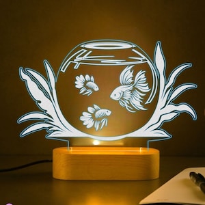 Könnte beinhalten: Ein weißes, 3D-gedrucktes Nachtlicht in Form einer Fischschale mit drei Fischen darin. Das Nachtlicht steht auf einem Holzsockel und hat ein warmweißes Leuchten. Das Bild zeigt auch zwei weitere Nachtlichter in verschiedenen Farben, gelb und lila.
