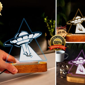 May include: A wooden base with a triangular acrylic panel featuring a white UFO with a person falling from it. The UFO is illuminated in blue, yellow, and purple in three separate images. The base is made of wood and has a natural finish.