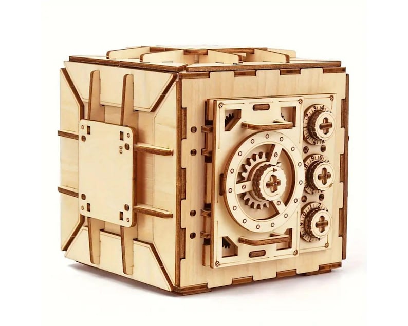 May include: A wooden model of a safe with a detailed, intricate design. The safe has a large dial with gears and a handle. The safe is made of light wood and has a natural finish.