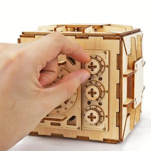 May include: A wooden model of a safe with three combination dials. The dials are set to 0, 0, and 0. A hand is turning the top dial.