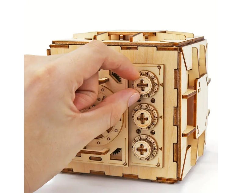 May include: A wooden model of a safe with three combination dials. The dials are set to 0, 0, and 0. A hand is turning the top dial.