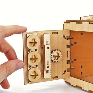 May include: A wooden model of a safe with a door that opens to reveal a small interior space. The door has a combination lock with three dials and a gear mechanism. The safe is made of light brown wood and has a natural finish.