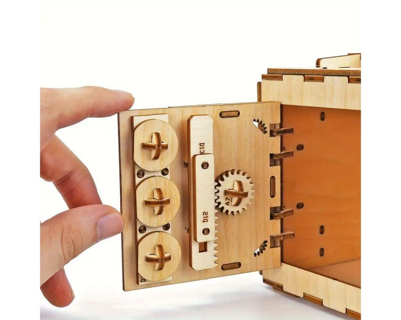 May include: A wooden model of a safe with a door that opens to reveal a small interior space. The door has a combination lock with three dials and a gear mechanism. The safe is made of light brown wood and has a natural finish.