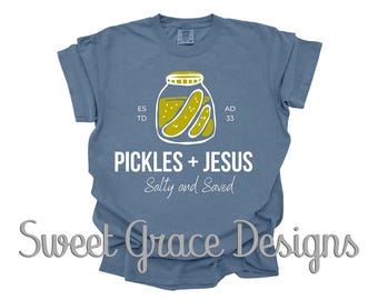 May include: A blue t-shirt with a graphic of a jar of pickles and the words "PICKLES + JESUS Salty and Saved". The design includes the text "ESTD AD 33". The shirt also has the text "Sweet Grace Designs".