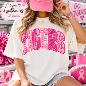 Op de afbeelding: Een gebroken wit t-shirt met het woord TIGERS in grote, roze letters in varsity-stijl. Elke letter heeft een uniek patroon, waaronder ruitjes, strepen, stippen en lintjes voor borstkankerbewustzijn in verschillende tinten roze.