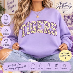 May include: A light purple crewneck sweatshirt featuring the word TIGERS in a collegiate font. The letters are filled with a yellow background and a repeating pattern of small purple roses. The sweatshirt has long sleeves and a ribbed neckline.