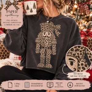 Può includere: Una felpa grigio antracite a maniche lunghe con una grande grafica di uno schiaccianoci con motivo leopardato. Lo schiaccianoci indossa un cappello abbinato con stampa leopardata e un fiocco. Il design presenta una texture grunge dettagliata.
