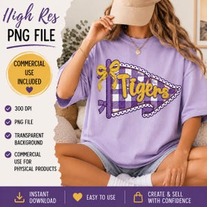 May include: A light purple t-shirt featuring a graphic of a purple and white checkered pennant flag with a yellow bow and the word Tigers in yellow script. The design includes a white scalloped border around the pennant shape.